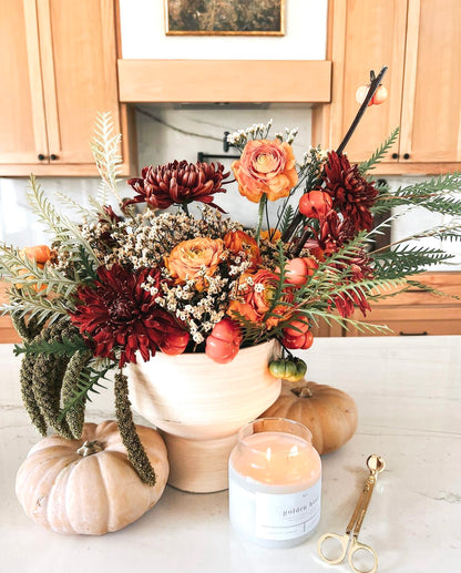 Golden Hour 100% soy wax candle lit beside a vibrant autumn floral arrangement, with white pumpkins and gold wick trimmer, creating a cozy, seasonal ambiance. Hand-poured, eco-friendly candle with notes of amber, lemon, ginger, patchouli, and wood, perfect for fall decor and clean, non-toxic home fragrance.