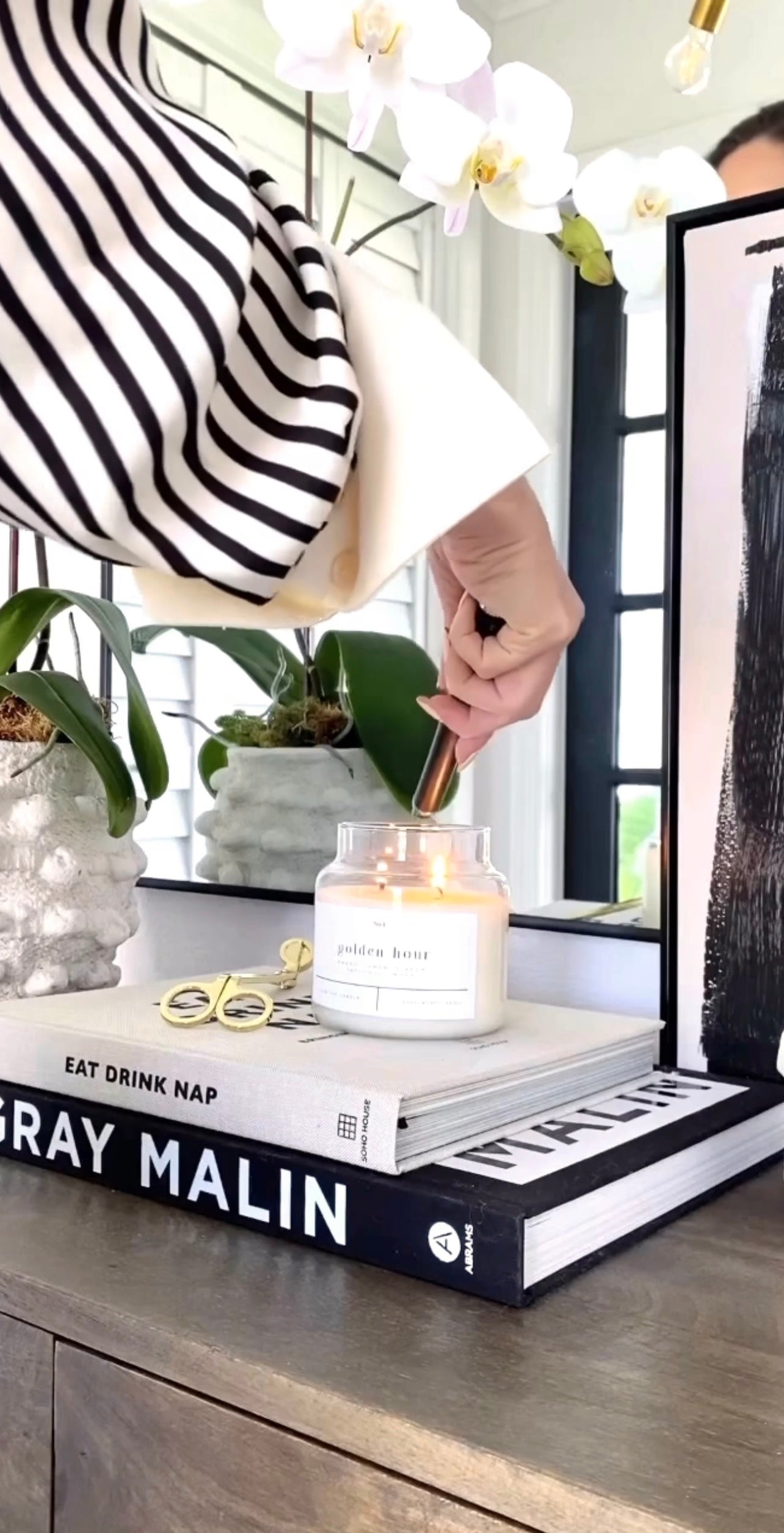Person trimming the wick of a lit Golden Hour 100% soy wax candle, styled on a modern coffee table with art books and a sleek orchid plant. Hand-poured, eco-friendly candle with a clean, non-toxic fragrance featuring notes of amber, lemon, ginger, patchouli, and wood.