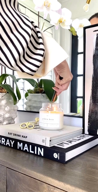 Person trimming the wick of a lit Golden Hour 100% soy wax candle, styled on a modern coffee table with art books and a sleek orchid plant. Hand-poured, eco-friendly candle with a clean, non-toxic fragrance featuring notes of amber, lemon, ginger, patchouli, and wood.